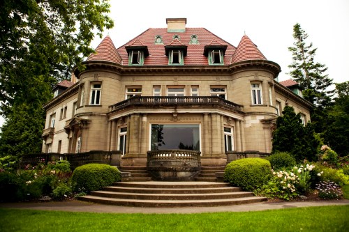 A grand, historic mansion with a stone facade, multiple windows, and lush greenery surrounding the entrance steps.