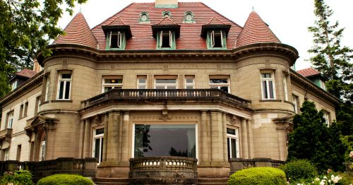 A grand, historic mansion with a large front staircase, surrounded by lush greenery and colorful flowers.