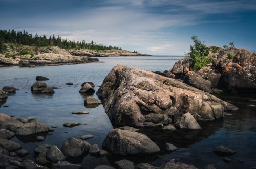 A serene coastal scene featuring large rocks and calm water, surrounded by trees under a cloudy sky.