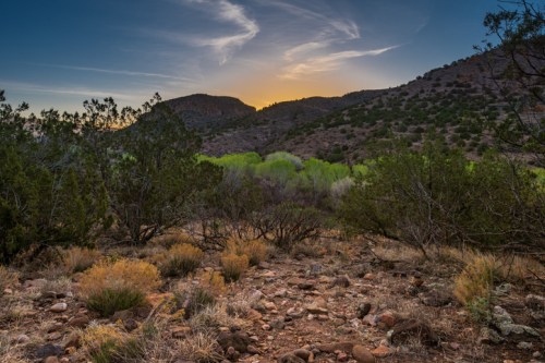 A scenic landscape at sunset, featuring rocky terrain, green foliage, and distant mountains under a colorful sky.