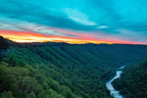 A vibrant sunset over a lush green valley with a winding river below, framed by rolling hills and a colorful sky.
