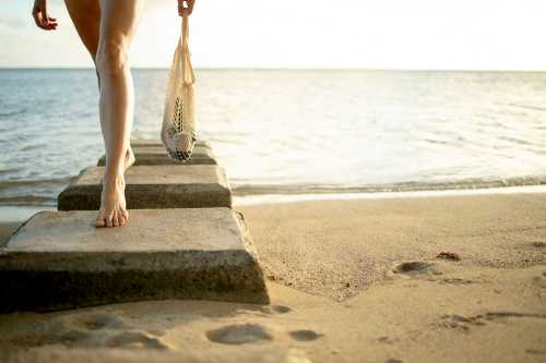 A person walks barefoot on stone steps leading to a beach, holding a net bag, with the ocean in the background.