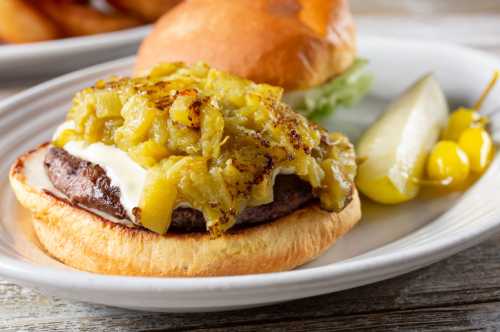 A close-up of a burger with a grilled patty, mayonnaise, and green chili topping, served with a lemon wedge and pickled peppers.