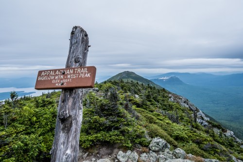 Signpost for the Appalachian Trail at Bigelow Mountain's West Peak, with a scenic view of mountains and cloudy sky.