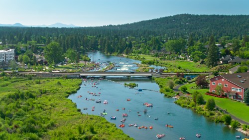 A scenic river with kayakers and lush greenery, surrounded by hills and buildings under a clear blue sky.