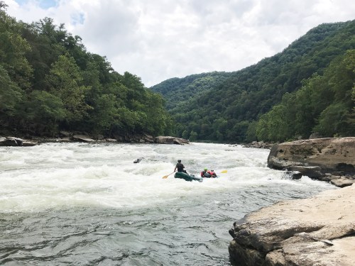Two kayakers navigate a turbulent river surrounded by lush green mountains under a cloudy sky.