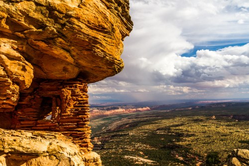 A rocky cliff with layered stone formations overlooks a vast green valley under a cloudy sky.