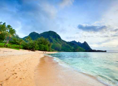 A serene beach with soft sand, gentle waves, and lush green mountains under a cloudy sky.