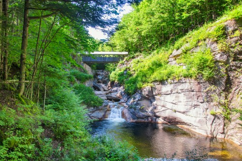 A serene landscape featuring a small waterfall, rocky terrain, and a bridge surrounded by lush green trees.