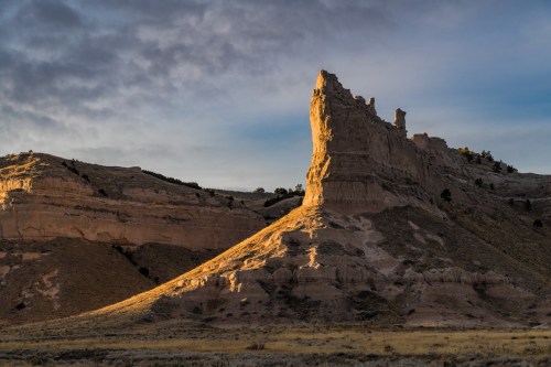 A dramatic rock formation rises against a cloudy sky, illuminated by warm sunlight in a rugged landscape.