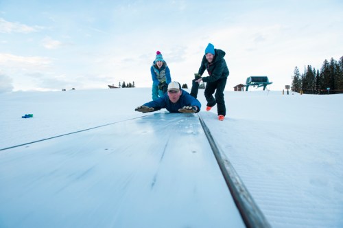 Three people play in the snow, with one lying on a slick surface while the others assist, set against a winter landscape.