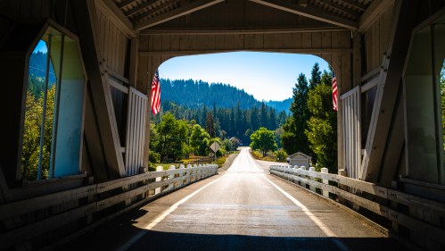View through a covered bridge, showcasing a scenic road lined with trees and mountains in the background.