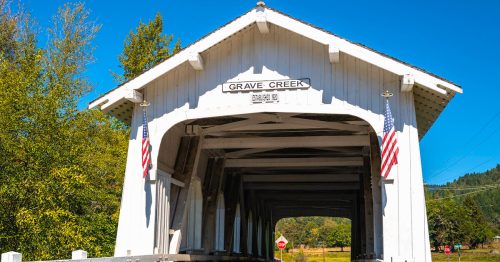 A white covered bridge with "Grave Creek" sign, flanked by trees and blue sky, inviting vehicles to cross.