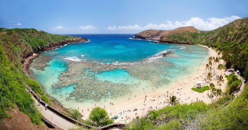 A panoramic view of a tropical beach with clear blue water, coral reefs, and lush green hills in the background.