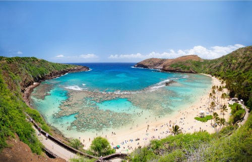 A panoramic view of a tropical beach with clear blue water, coral reefs, and lush green hills in the background.