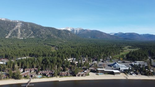 Aerial view of a lakeside town surrounded by mountains and dense forests under a clear blue sky.