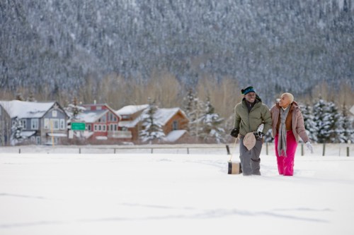 A couple walks through a snowy landscape, smiling, with houses and mountains in the background.