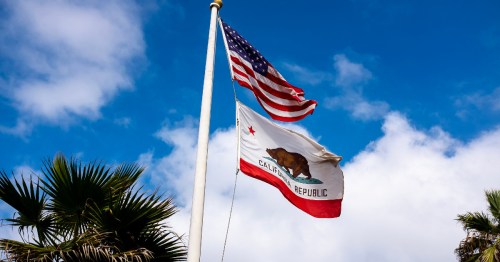 Flags of the United States and California waving against a blue sky with clouds, surrounded by palm trees.