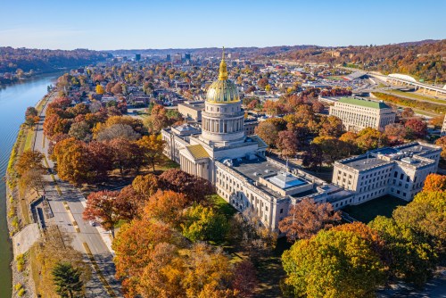 Aerial view of a state capitol building surrounded by autumn foliage and a river, with a city skyline in the background.
