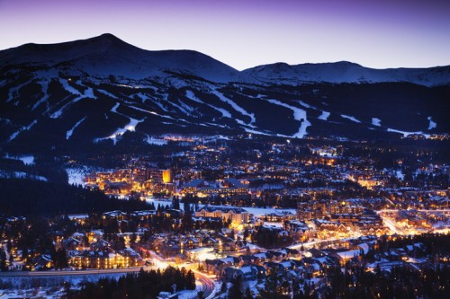 A snowy mountain town at dusk, with twinkling lights and ski slopes in the background under a purple sky.