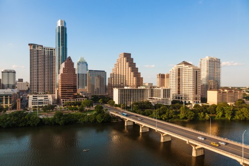 Aerial view of a city skyline with modern skyscrapers beside a river and a bridge, under a clear blue sky.