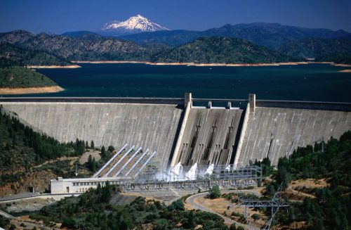 Aerial view of a large dam with water flowing, surrounded by mountains and a lake under a clear blue sky.