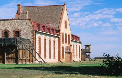 Historic stone building with a red roof, surrounded by green grass and a watchtower in the background under a blue sky.