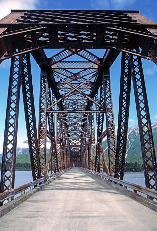 A view of a rusted metal bridge with intricate latticework, spanning over a river against a backdrop of mountains and blue sky.