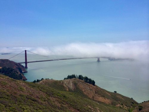 Golden Gate Bridge partially shrouded in fog, spanning a bay with green hills in the foreground and clear blue sky above.