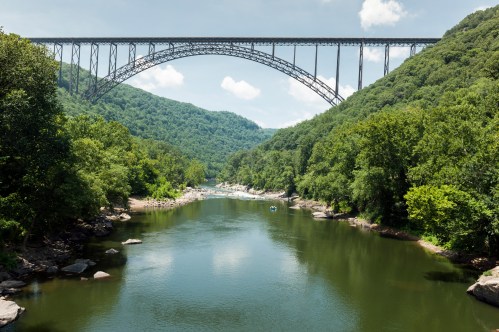 A scenic view of a river surrounded by lush greenery, with a large bridge arching over it under a clear blue sky.