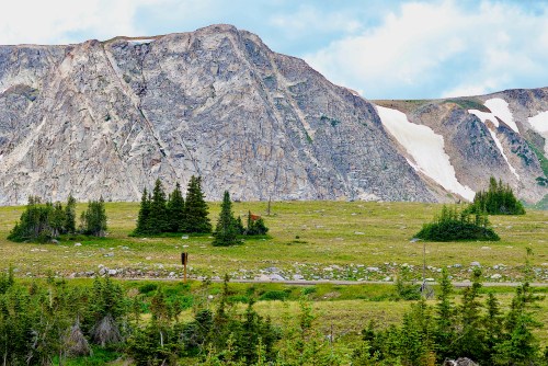 A rugged mountain landscape with rocky cliffs, patches of snow, and green vegetation in the foreground.
