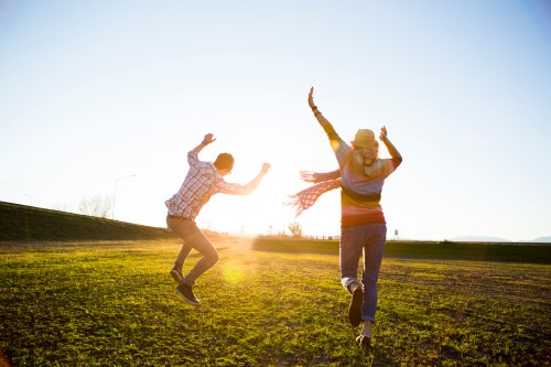 Two people joyfully jumping in a sunlit field, with arms raised and a clear blue sky in the background.