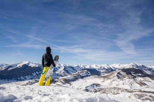 A person in a black jacket and yellow pants stands on a snowy mountain, gazing at a panoramic view of snow-capped peaks.