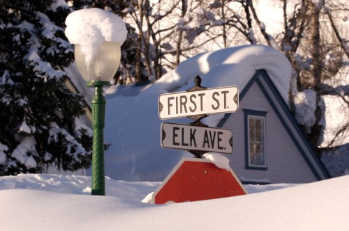 Snow-covered street signs for First St. and Elk Ave. near a lamp post, with a house in the background.