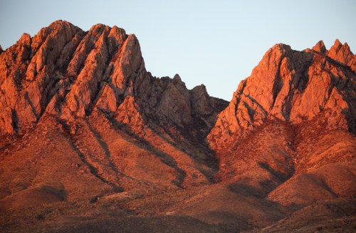 Sunlit rocky mountains with rugged peaks and warm hues, set against a clear sky.