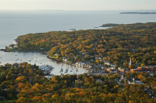 Aerial view of a coastal town surrounded by autumn foliage, with boats in the harbor and a calm sea in the background.