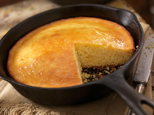 A golden cornbread in a cast iron skillet, with a slice cut out, resting on a textured cloth.