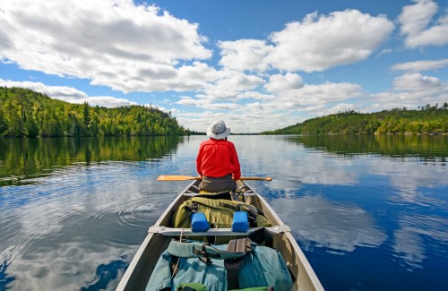 A person in a red shirt sits in a canoe, paddling on a calm lake surrounded by lush greenery and blue skies.