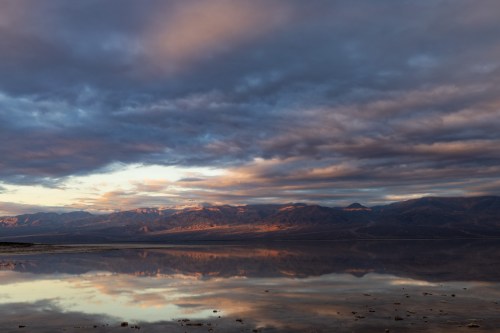 A serene landscape with mountains reflecting in calm water under a dramatic sky filled with clouds at sunset.