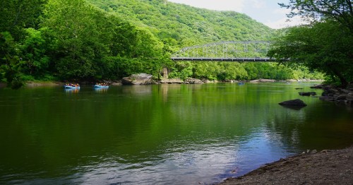A serene river scene with a bridge, surrounded by lush greenery and people rafting in the water.