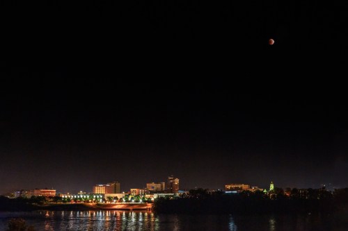 A city skyline at night with a river, illuminated buildings, and a partial lunar eclipse in the sky.