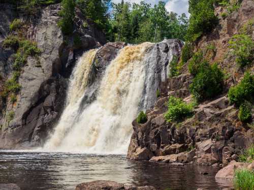 A cascading waterfall flows over rocky cliffs, surrounded by lush greenery and a calm river below.