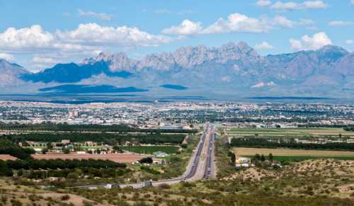 A scenic view of a highway leading to a city, with mountains in the background and lush fields in the foreground.