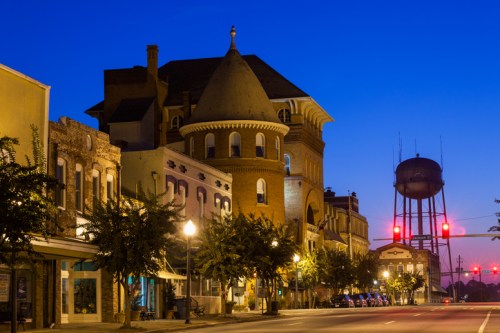 Historic buildings line a quiet street at dusk, with a water tower visible in the background under a blue sky.