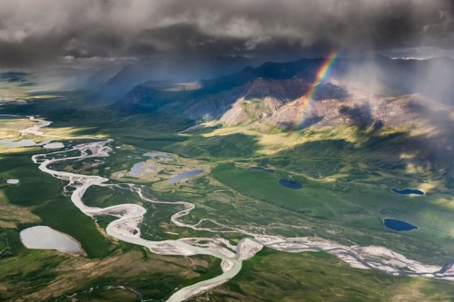 Aerial view of a lush landscape with winding rivers, lakes, and a rainbow against a backdrop of mountains and clouds.