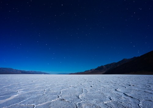 A vast, cracked salt flat under a starry night sky, with distant mountains silhouetted against the horizon.