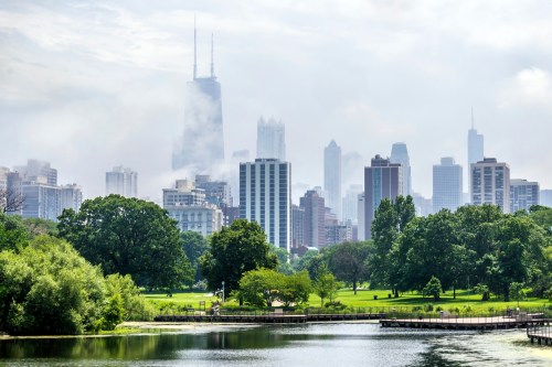 A lush green park with a lake, framed by a skyline of tall buildings and a foggy sky in the background.