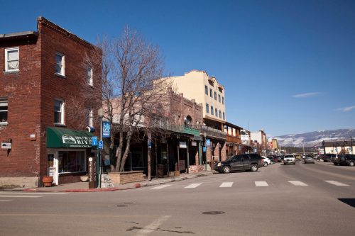 A charming street scene featuring brick buildings, shops, and parked cars under a clear blue sky. Mountains in the background.