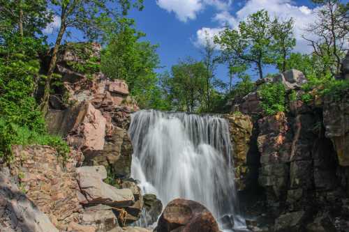 A serene waterfall cascades over rocky cliffs, surrounded by lush greenery and a bright blue sky.