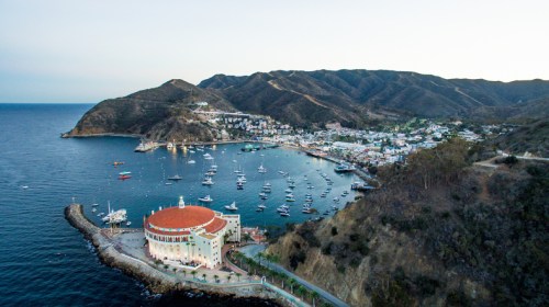 Aerial view of a coastal town with a harbor, boats, and mountains in the background during sunset.